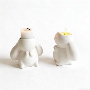 May include: Two white ceramic bunny candle holders. One holds a lit white candle, the other a lit tea light. The bunny figures have large ears and a bow detail. The candle holders are on a white background.