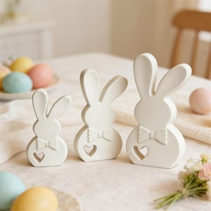 May include: Three white ceramic Easter bunny ornaments in different sizes. Each bunny has long ears, a bow tie, and a heart-shaped cutout. They are displayed on a cream tablecloth, with pastel Easter eggs visible in the background.