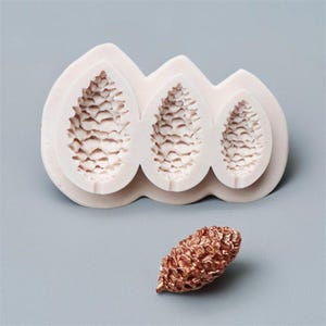 May include: A white silicone mould featuring three pine cone impressions of different sizes. A separate, small, bronze-coloured pine cone is also present. The mould is designed for crafting or baking, with detailed textures for realistic results.