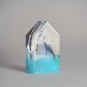 Op de afbeelding: Een transparant glazen sculptuur van twee walvissen die uit het water springen. Het sculptuur heeft de vorm van een huis met een blauwe basis en een wit en grijs interieur. De walvissen zijn geschilderd in tinten blauw en grijs.