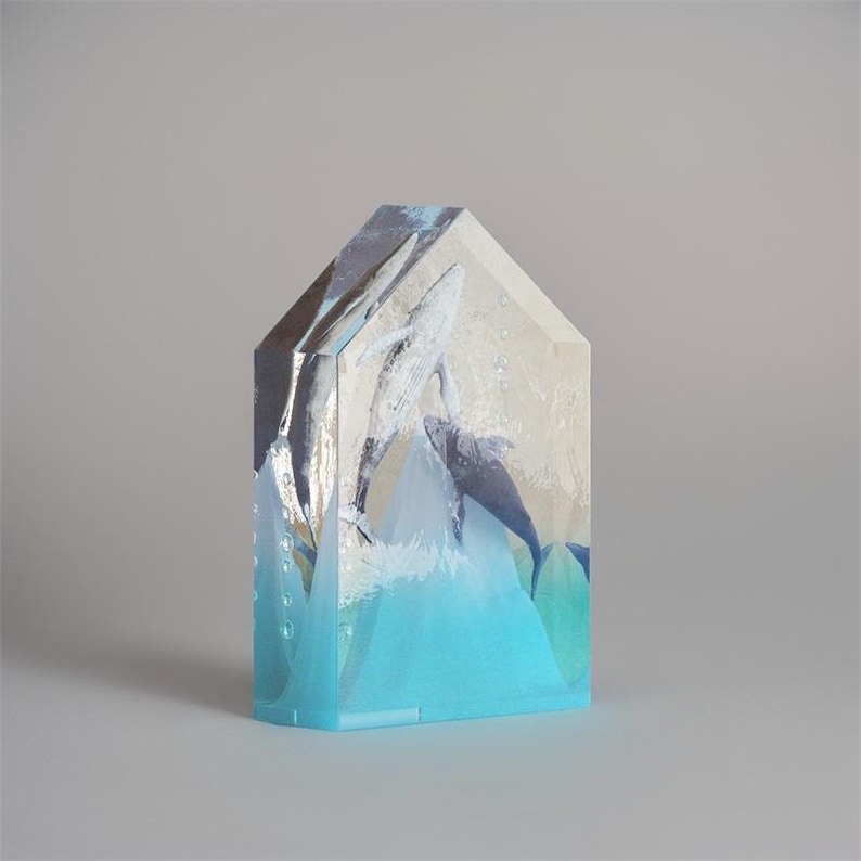 Op de afbeelding: Een transparant glazen sculptuur van twee walvissen die uit het water springen. Het sculptuur heeft de vorm van een huis met een blauwe basis en een wit en grijs interieur. De walvissen zijn geschilderd in tinten blauw en grijs.