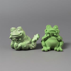 Könnte beinhalten: Zwei grüne Froschfiguren. Ein Frosch liegt mit ausgestrecktem Arm, der andere sitzt aufrecht mit den Händen am Gesicht. Beide Frösche haben eine strukturierte Oberfläche und sind in einer spielerischen Pose.