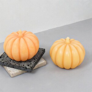 May include: Two small, orange pumpkin-shaped candles on a grey surface. The candles are made of wax and have a ribbed texture.