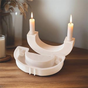 May include: Two white, modern candle holders with lit candles. The holders have a curved design with a square base for the candles. A matching, unlit holder is in the foreground. The candles are white and cylindrical.