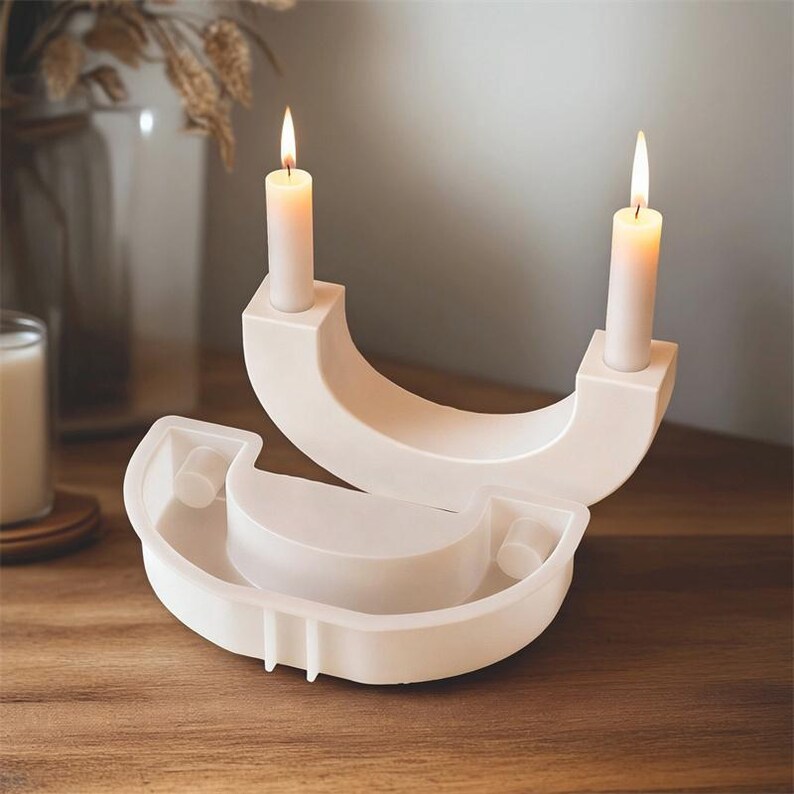 May include: Two white, modern candle holders with lit candles. The holders have a curved design with a square base for the candles. A matching, unlit holder is in the foreground. The candles are white and cylindrical.