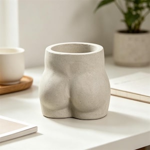 Op de afbeelding: Een lichtgrijze, gespikkelde betonnen plantenbak in de vorm van een menselijke torso. De pot heeft een rond, sculpturaal ontwerp met een brede cirkelvormige opening aan de bovenkant en een platte basis, gemaakt van mat, getextureerd materiaal.