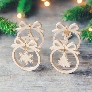 Puede incluir: Cuatro adornos navideños color crema con detalles de lazo. Cada adorno presenta un diseño recortado diferente: una estrella, un copo de nieve, un ciervo y un árbol de Navidad. Los adornos están suspendidos por cadenas doradas.