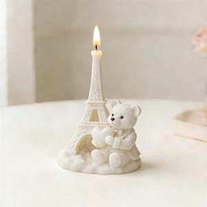 May include: A cream-coloured candle in the shape of the Eiffel Tower, with a lit wick. A white teddy bear holding a heart sits in front. The candle and bear are on a cloud-shaped base.