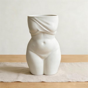 Puede incluir: Un jarrón de cerámica blanca con forma de torso femenino. El jarrón presenta una parte superior drapeada, que imita un corpiño, y una forma suave y curvilínea. El diseño es minimalista y moderno, adecuado para la decoración del hogar.
