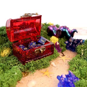 May include: A red glitter treasure chest with a brass latch is open and filled with blue dice with gold numbers. The chest is sitting on a bed of green moss with yellow and purple crystals scattered around it.