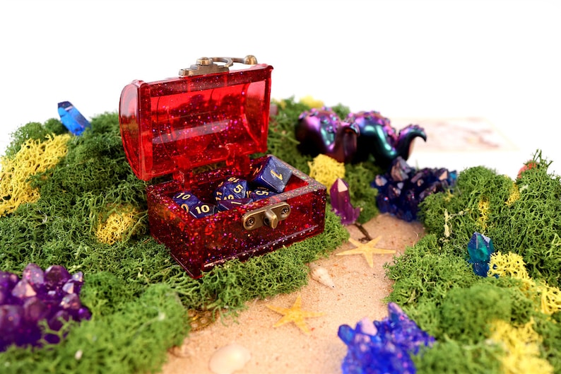 May include: A red glitter treasure chest with a brass latch is open and filled with blue dice with gold numbers. The chest is sitting on a bed of green moss with yellow and purple crystals scattered around it.