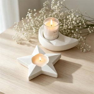 Puede incluir: Una vela encendida en un vaso transparente se encuentra en un soporte de cerámica blanca en forma de media luna. Un portavelas blanco en forma de estrella con una vela encendida está en primer plano. Flores blancas en el fondo.