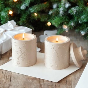 Puede incluir: Dos velas beige encendidas en soportes cilíndricos con diseños de copos de nieve y árboles de Navidad. Una vela tiene la tapa quitada. Las velas están sobre una superficie blanca, con un árbol de Navidad y una caja de regalo en el fondo.