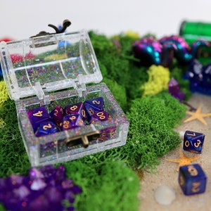 May include: A clear plastic treasure chest filled with purple and blue dice with gold numbers. The chest is sitting on a bed of green moss with other purple and blue dice scattered around it.