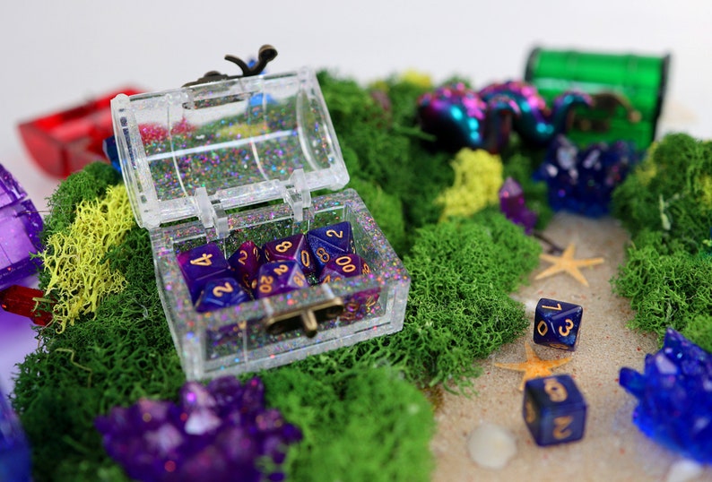 May include: A clear plastic treasure chest filled with purple and blue dice with gold numbers. The chest is sitting on a bed of green moss with other purple and blue dice scattered around it.