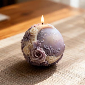 May include: A spherical candle with a lit wick, featuring a detailed floral design. The candle is a soft lavender color, adorned with sculpted roses and other flowers in shades of purple and cream. The candle is approximately 4 inches in diameter.