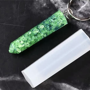 May include: A clear silicone mold for making resin keychains. The mold is shaped like a long, thin rectangle with rounded edges. A finished keychain made with the mold is shown, featuring green glitter embedded in clear resin.