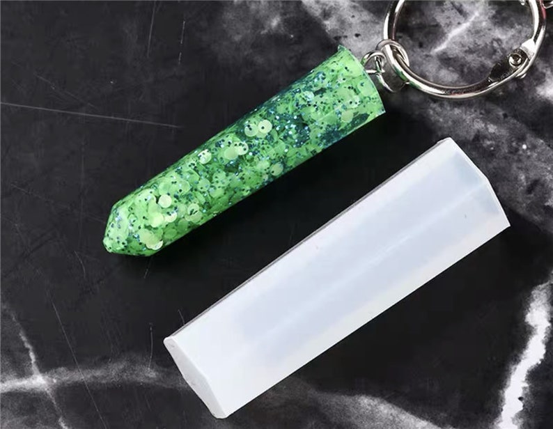 May include: A clear silicone mold for making resin keychains. The mold is shaped like a long, thin rectangle with rounded edges. A finished keychain made with the mold is shown, featuring green glitter embedded in clear resin.