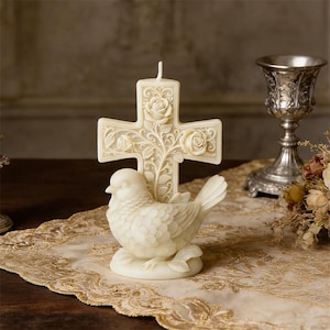 May include: A cream-colored candle shaped like a cross with rose and vine detailing, standing behind a detailed bird figurine. The candle and bird are on a gold-colored, patterned surface, with a silver chalice in the background.