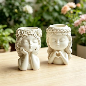 May include: Two off-white ceramic planters shaped like girls with closed eyes. One is blowing a kiss, the other is making a heart shape with her hands. Both have floral crowns and are on a light brown surface.