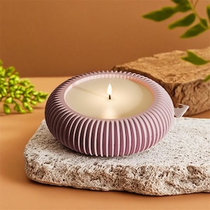 Könnte beinhalten: Eine brennende, runde Kerze in einem gerippten Behälter in Malvenfarbe. Die Kerze steht auf einem strukturierten, hellgrauen Stein. Der Hintergrund ist ein warmes Braun mit einem Stück braunem Stein und grünem Laub.