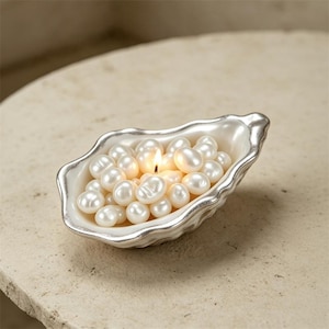 May include: A decorative oyster-shaped dish with a metallic silver rim, filled with numerous white, lustrous, spherical pearls. A small, lit candle flame emerges from the center of the pearl cluster, casting a warm glow on the surrounding white beads.