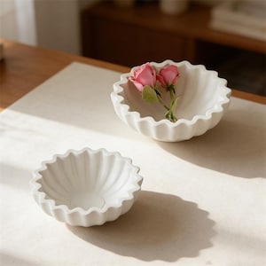 May include: Two white ceramic bowls with a scalloped edge. One bowl contains two pink roses. The bowls are on a cream-colored surface, with a wooden table and cabinet in the background. The bowls have a fluted design.