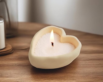 Puede incluir: Una vela en forma de corazón con un exterior color crema y una mecha encendida. El interior de la vela está lleno de gránulos blancos. La vela está sobre una superficie de madera.