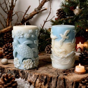 May include: Two decorative pillar candles with ocean-themed designs. One candle features a sea turtle and flowers, the other depicts birds and waves. The candles are blue and white, set on a wooden surface with pine cones.