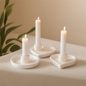 Puede incluir: Tres candelabros de cerámica blanca con velas blancas encendidas. Los candelabros tienen forma de corazón y redondos, colocados sobre una superficie de color neutro. Las velas son cilíndricas y brillan suavemente.