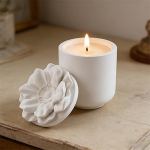 May include: A white cylindrical candle in a matte container with a lit wick. Beside the candle sits a matching circular lid featuring a three-dimensional sculpted flower design on its top surface. The candle emits a warm, soft glow.