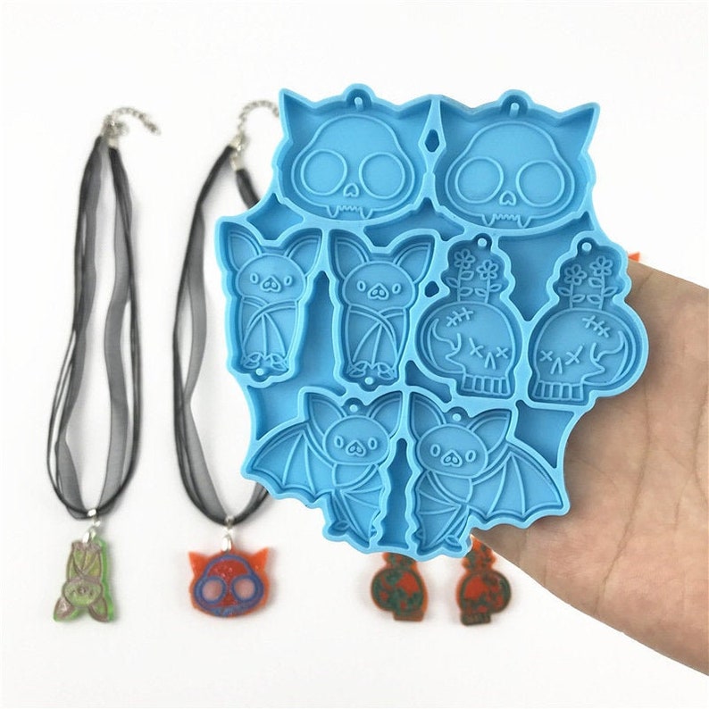 Puede incluir: Un molde de silicona azul con ocho formas diferentes de tem&aacute;tica de Halloween, incluyendo murci&eacute;lagos, calaveras y una planta. El molde est&aacute; en una mano, y hay varias piezas de resina terminadas al lado.