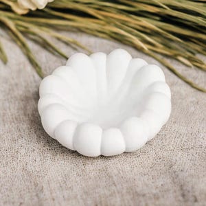 May include: A white, scalloped-edge ceramic dish, resembling a seashell. The dish is round with a shallow interior and a textured surface. The background features dried foliage on a neutral linen cloth.