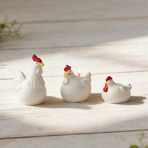 May include: Three white ceramic chicken figurines with red combs and yellow beaks. One chicken has a candle lit inside. The chickens are arranged on a light-colored wooden surface, creating a charming and rustic aesthetic.