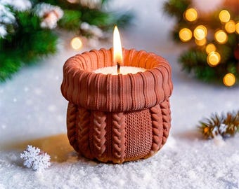 Puede incluir: Una vela encendida en un portavelas de color terracota, estilo suéter. La vela tiene un interior de cera blanca y una larga llama parpadeante. El soporte tiene una parte superior acanalada y un diseño de punto de cable en el cuerpo. La escena está sobre una superficie nevada.