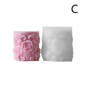 May include: A pink candle with a detailed rose design and a white mold. The candle features a central rose surrounded by leaves and smaller roses. The mold is a simple, smooth shape.