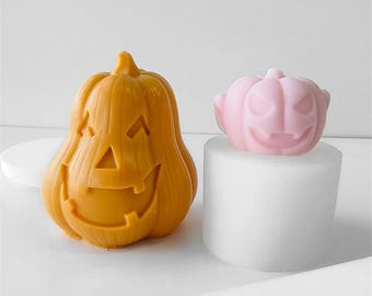 Könnte beinhalten: Zwei Halloween-kürbisförmige Kerzen, eine orange und eine rosa, mit geschnitzten Jack-o'-Lantern-Gesichtern. Die Kerzen stehen auf weißen Sockeln.