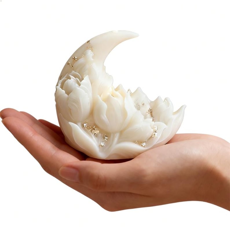 May include: A white, crescent moon-shaped candle with floral detailing. The candle is decorated with gold flecks and held in a hand. The candle is a decorative item.