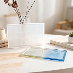 May include: A rectangular silicone mould with a grid pattern stands upright, while a finished rectangular tray with a smooth surface and a gradient of yellow and blue resin sits flat in front of it, showcasing a clear, glossy finish.