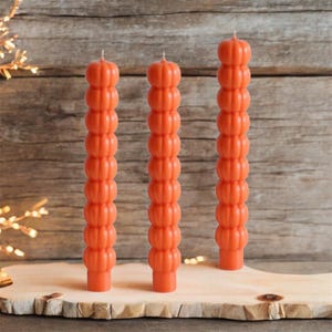 May include: Three orange pillar candles with a pumpkin-like design. The candles are standing upright on a wooden surface against a rustic wooden background. The candles are a vibrant orange color and have a textured, segmented appearance.