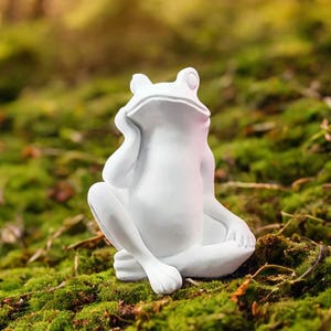 Puede incluir: Una figura de rana blanca en posición sentada, apoyando la barbilla en la mano. La rana se encuentra sobre musgo verde, creando una escena natural. La figura tiene un acabado liso y mate y es un adorno decorativo para jardín.