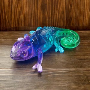 May include: A translucent chameleon figurine in shades of purple, blue, and green. The sculpture has a curled tail and detailed features, resting on a wooden surface. The chameleon's body transitions from purple to blue, with a green tail.