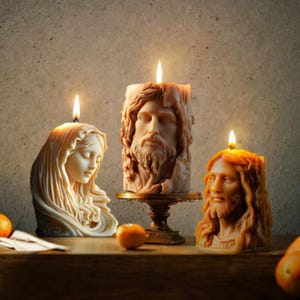 May include: Three religious-themed candles with lit wicks. One candle depicts a woman's face, another a man's face, and the third a man's face. The candles are various shades of beige and brown, with a wooden surface in the foreground.