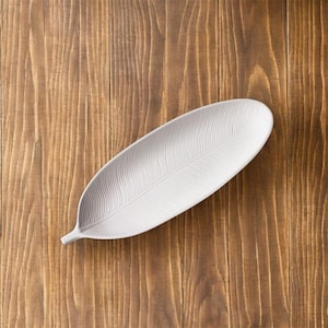 Può includere: Un piatto da portata in ceramica bianca a forma di foglia con un motivo a piuma strutturato. Il piatto è posto su una superficie di legno. Il piatto è lungo circa 36 cm.