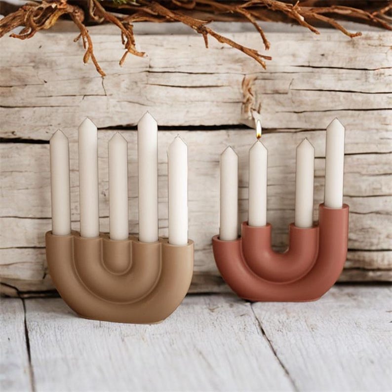 May include: Two modern candle holders, one tan and one terracotta, each holding six white candles. One candle is lit. The holders have a curved, arch-like design. The background is a rustic wooden surface.