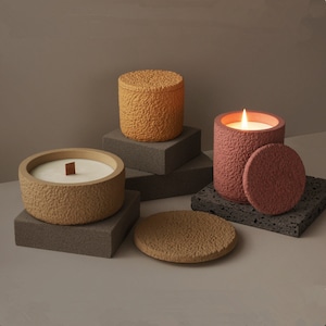 Peut inclure: Trois pots de bougies texturés avec des couvercles. Les bougies sont dans des tons de brun et de rose. Les bougies sont sur une surface texturée grise.