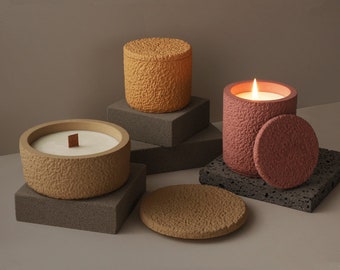 May include: Three textured candle jars with lids. The candles are in shades of brown and pink. The candles are on a gray textured surface.