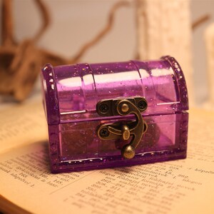 May include: A small purple plastic treasure chest with a gold-toned latch and hinges. The chest is sitting on a book with text.