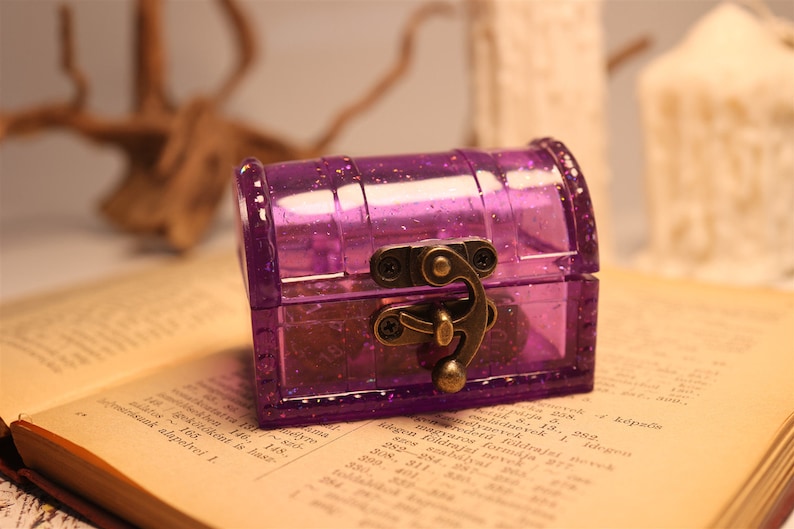 May include: A small purple plastic treasure chest with a gold-toned latch and hinges. The chest is sitting on a book with text.