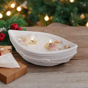 Op de afbeelding: Een witte keramische bootvormige kandelaar met twee brandende kaarsen, schelpen en imitatieparels. De houder staat op een houten oppervlak, met een kerstboom en lichten op de achtergrond. De kandelaar is versierd met een touwontwerp.